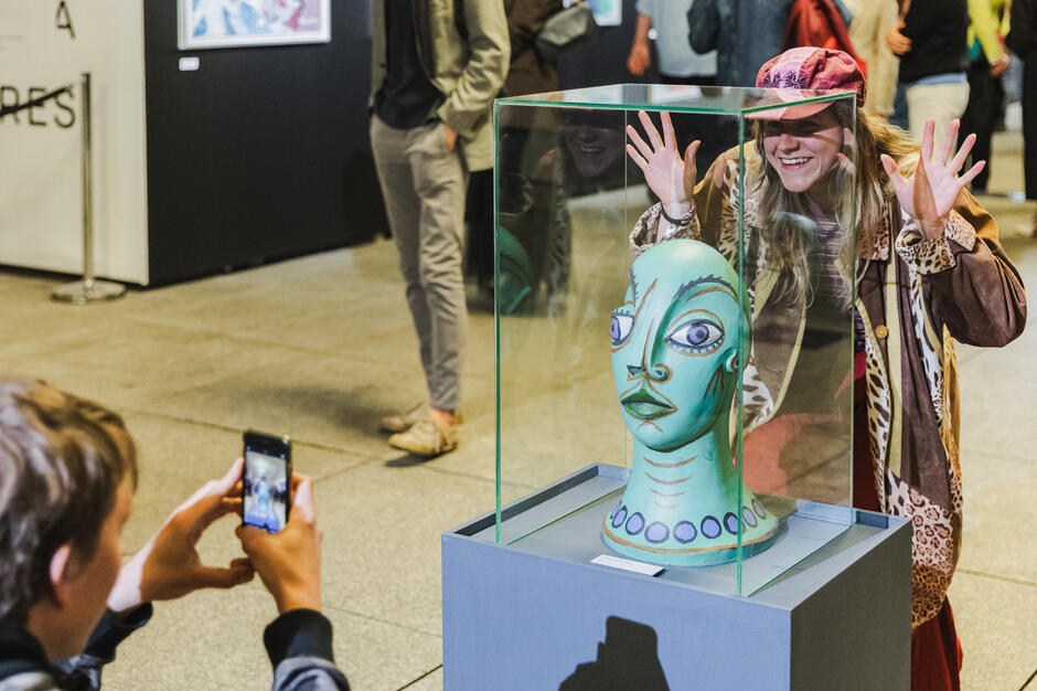 Kobieta z szerokim uśmiechem pozuje za szklaną gablotą, w której znajduje się rzeźba kolorowej głowy o fantazyjnych rysach twarzy. Chłopiec przed nią robi zdjęcie telefonem, uchwytując moment radości i zainteresowania.