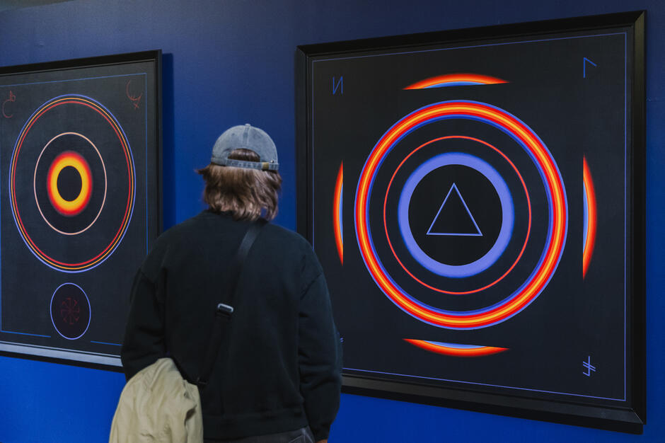 Młody mężczyzna w czapce z daszkiem ogląda intensywnie nowoczesne obrazy z koncentrycznymi kręgami i symbolami geometrycznymi na ciemnym tle. Dzieła charakteryzują się wyrazistymi barwami i aurą tajemniczości, przyciągając wzrok swoją precyzją.