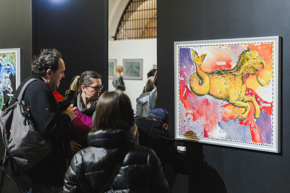 Kilka osób z bliska ogląda barwny obraz przedstawiający mityczną bestię przypominającą lwa z rybim ogonem, unoszącego się na tle kolorowych map i nazw gwiazdozbiorów. Jeden z widzów przyklęka, aby przeczytać podpis pod dziełem, co świadczy o głębokim zainteresowaniu jego treścią.