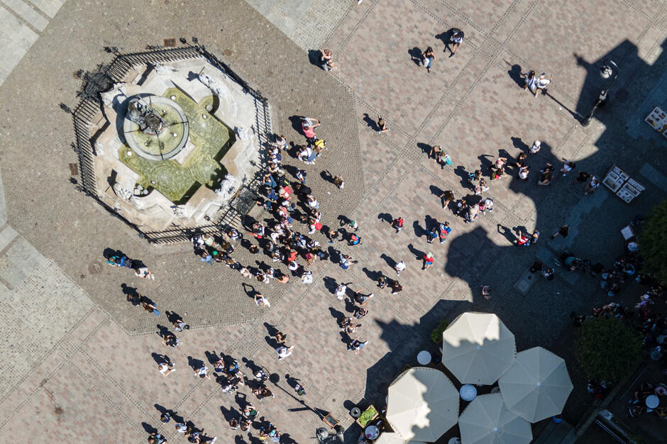 Zdjęcie z lotu ptaka ukazuje tłum ludzi zgromadzonych wokół zabytkowej fontanny na brukowanym placu, otoczonej ozdobną balustradą. W prawym dolnym rogu widoczne są parasole ogródka kawiarnianego, a cień pobliskiego budynku nadaje kompozycji charakterystyczny, graficzny kształt.