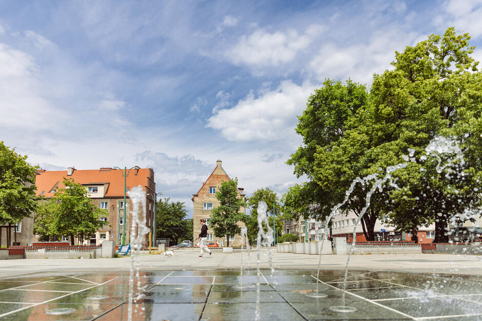 Na zdjęciu widać nowoczesną fontannę posadzkową na miejskim placu, z której woda tryska w rytmicznych łukach, tworząc dynamiczną kompozycję. W tle znajdują się drzewa oraz budynki mieszkalne z czerwonymi dachami, a przez plac przechodzi kobieta z psem, co dodaje scenie codziennego, spokojnego charakteru.