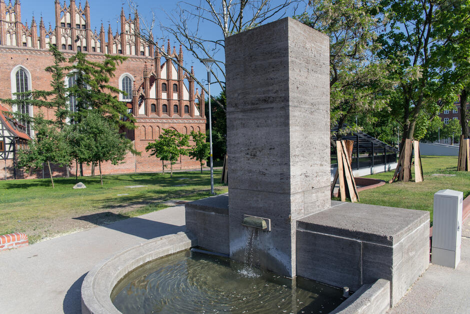 Na zdjęciu znajduje się nowoczesna fontanna o prostej, geometrycznej formie, z której woda wypływa z betonowego bloku do okrągłego basenu. W tle widoczny jest imponujący, gotycki kościół z charakterystycznymi ceglanymi elewacjami i bogato zdobionymi szczytami, otoczony zielenią i drzewami.