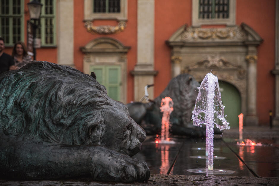 Na zdjęciu widoczna jest fontanna z kolorowym oświetleniem, otoczona rzeźbami leżących lwów z brązu, które nadają miejscu majestatyczny charakter. W tle znajduje się zabytkowy budynek z ozdobnymi portalami i pastelowymi okiennicami, co podkreśla historyczny klimat otoczenia.