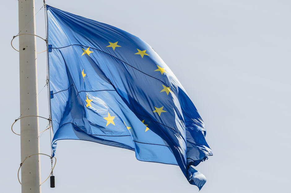 Wielka flaga Unii Europejskiej powiewa na potężnym maszcie, na tle niebieskiego słonecznego nieba