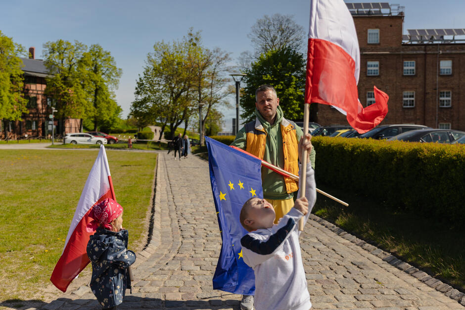 Ojciec na spacerze z dwoma małymi chłopcami, którzy trzymają w rękach flagi dwie biało-czerwone i jedną europejską