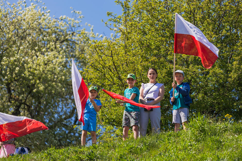 Kobieta stoi na zielonym od trawy wzgórzu, po jej obu stronach są mali chłopcy z biało-czerwonymi flagami 