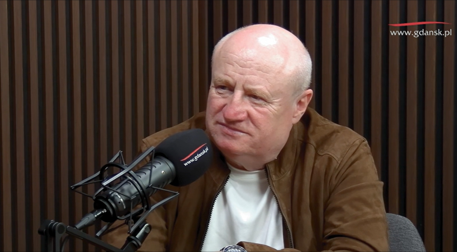 Na zdjęciu widać starszego mężczyznę w studiu nagraniowym, siedzącego przy mikrofonie z logo „www.gdansk.pl”. Ubrany jest w białą koszulkę i brązową kurtkę zamszową, a za nim znajduje się tło z pionowych, drewnianych paneli akustycznych