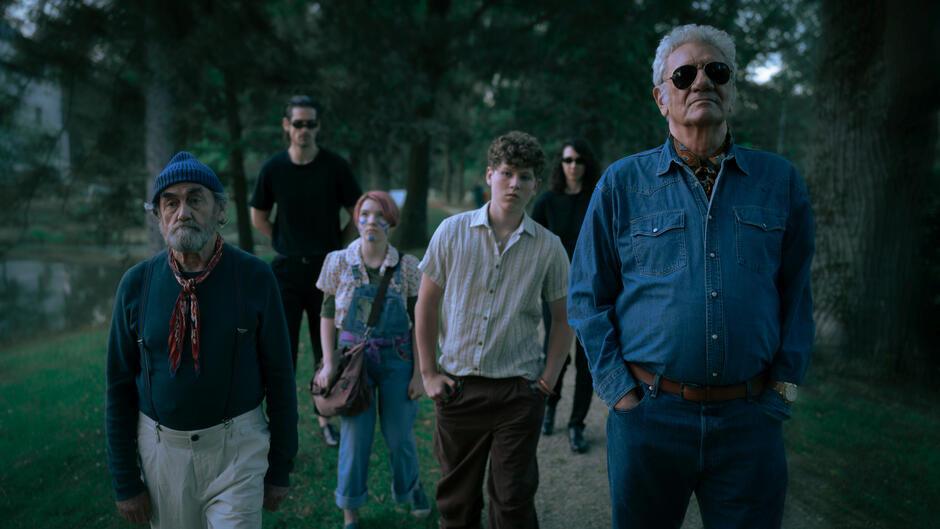 Grupa bohaterów w różnym wieku idzie przez las w filmowym, dramatycznym nastroju – na czele starszy mężczyzna w dżinsowej koszuli i ciemnych okularach. Wszyscy patrzą poważnie przed siebie, co buduje atmosferę tajemniczej misji lub ucieczki