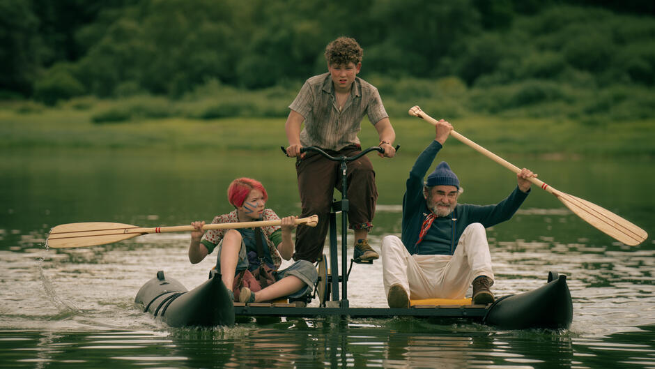 Trójka bohaterów płynie przez jezioro na nietypowej łodzi napędzanej pedałami i wiosłami – starszy pan, dziewczynka z różowymi włosami i chłopiec skupiony na kierowaniu. Ich determinacja i ekspresja wskazują na przygodę lub ucieczkę