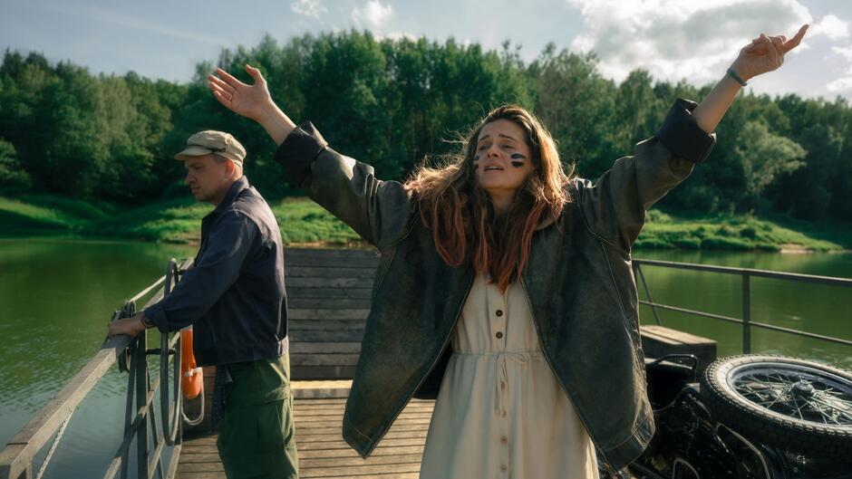 Kobieta z długimi włosami z różowymi końcówkami, w rozpiętej kurtce i pomalowaną twarzą, stoi na promie z uniesionymi ramionami w geście triumfu lub wolności. Obok niej mężczyzna spogląda w wodę, a w tle widać zieleń i spokój rzeki