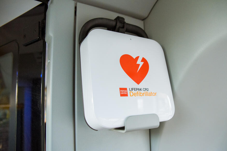 Na zdjęciu widoczny jest defibrylator LIFEPAK CR2 marki Physio-Control, zamontowany w specjalnym uchwycie wewnątrz pojazdu. Urządzenie jest oznaczone wyraźnym symbolem serca z błyskawicą, co sygnalizuje jego przeznaczenie do ratowania życia w przypadku nagłego zatrzymania krążenia.