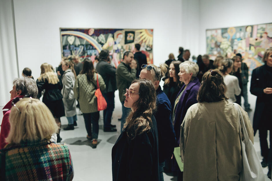 Duża grupa ludzi w galerii, z tyłu na ścianach wielkoformatowe kolorowe gobeliny