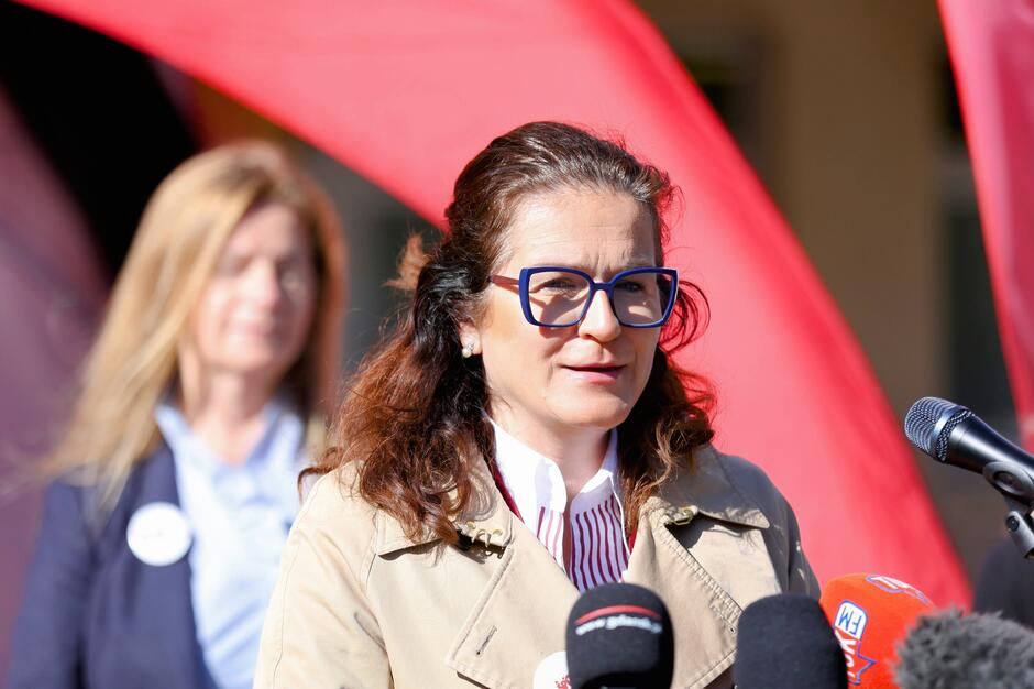 Na zdjęciu widać kobietę przemawiającą do mediów. Stoi przy mikrofonach oznaczonych logotypami różnych stacji radiowych i telewizyjnych, co sugeruje, że odbywa się konferencja prasowa lub publiczne wystąpienie. Kobieta ma na sobie beżowy płaszcz i okulary w niebieskich oprawkach. W tle widać czerwoną tkaninę oraz drugą kobietę, nieco rozmazaną, co wskazuje na głębię ostrości skupioną na osobie pierwszoplanowej.