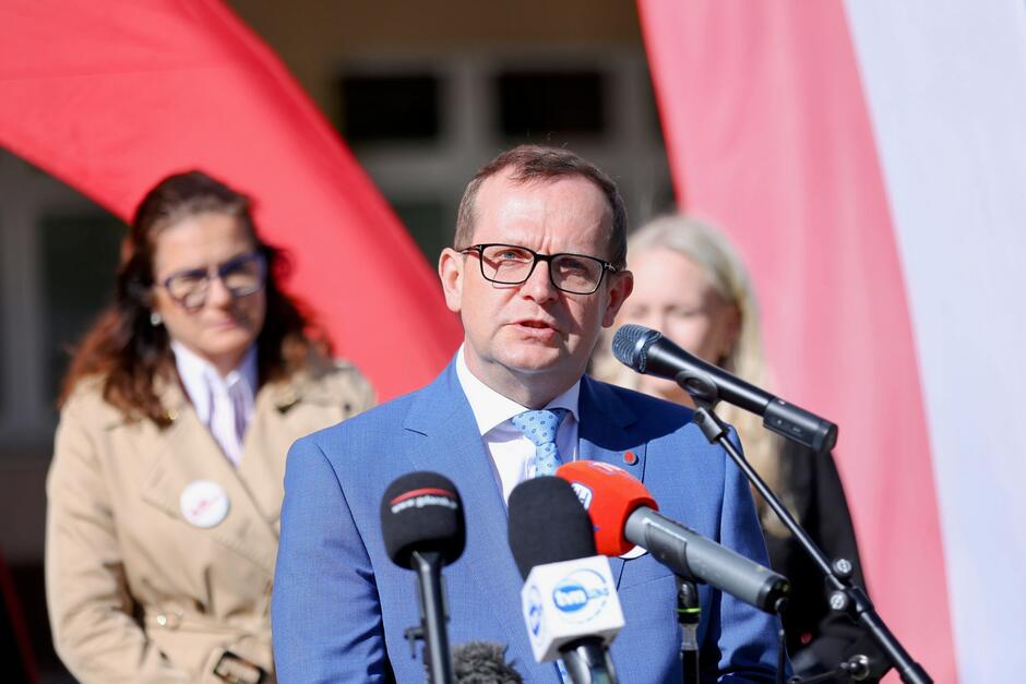 Na zdjęciu widać mężczyznę przemawiającego podczas konferencji prasowej. Ubrany jest w niebieski garnitur i jasnoniebieski krawat w kropki. Stoi przed mikrofonami różnych mediów, m.in. TVN24 i Radia Gdańsk, co sugeruje, że wydarzenie jest relacjonowane przez ogólnopolskie media. W tle widoczne są osoby, w tym kobieta w beżowym płaszczu — ta sama, która pojawiła się na poprzednim zdjęciu. Całość odbywa się na tle dużej flagi Polski i czerwonych materiałów, co wskazuje na oficjalny charakter wydarzenia, prawdopodobnie związany z kampanią wyborczą lub promocją udziału w wyborach.