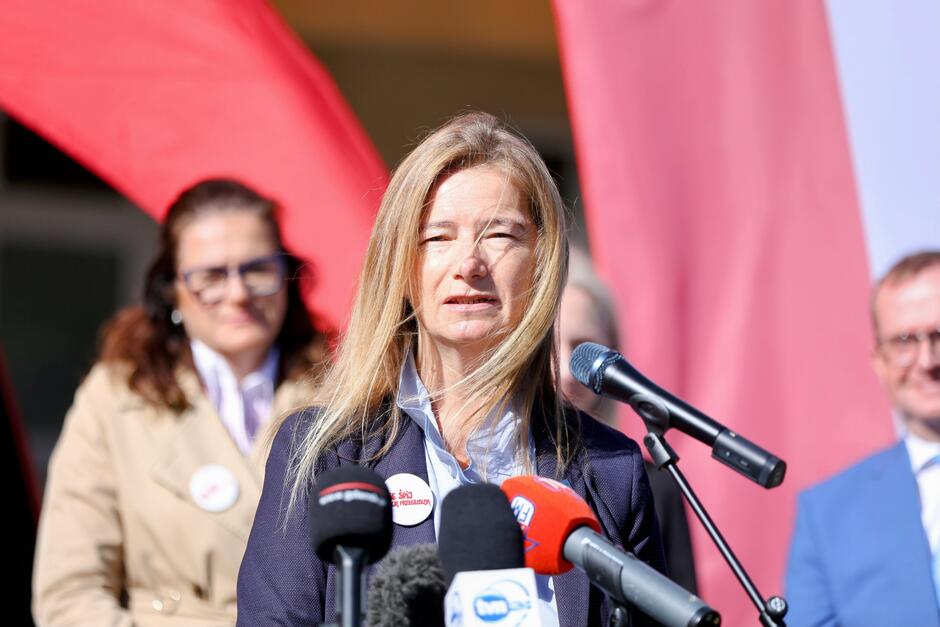 Na zdjęciu widać kobietę przemawiającą do mikrofonów podczas oficjalnego wydarzenia lub konferencji prasowej. Ubrana jest w granatową marynarkę, a na jej piersi widoczna jest przypinka z napisem „Zmień kraj. Idź na wybory” lub podobnym hasłem wyborczym (tekst nie jest całkowicie czytelny, ale związany z mobilizacją do udziału w wyborach). W tle znajdują się dwie inne osoby znane z poprzednich zdjęć — kobieta w beżowym płaszczu i mężczyzna w niebieskim garniturze — co wskazuje, że wszystkie te fotografie pochodzą z jednej konferencji lub wydarzenia promującego udział w wyborach prezydenckich 2025 w Polsce.