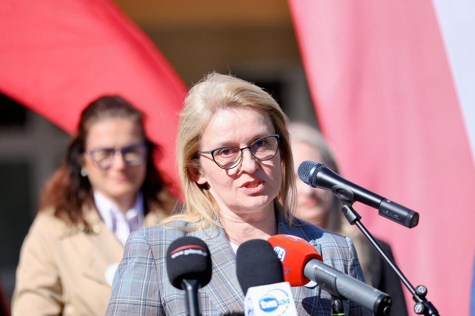 Na tym zdjęciu widzimy kolejną kobietę przemawiającą podczas tej samej konferencji prasowej lub wydarzenia związanego z wyborami prezydenckimi w Polsce, które – jak wynika z wcześniejszych materiałów – odbędą się 18 maja 2025 roku. Kobieta ma jasne włosy, okulary i nosi elegancką marynarkę w kratę. Przemawia do zebranych dziennikarzy, przed mikrofonami stacji takich jak TVN24, Radio Gdańsk i RMF FM. W tle znów widoczna jest kobieta w beżowym płaszczu, co potwierdza ciągłość wydarzenia.