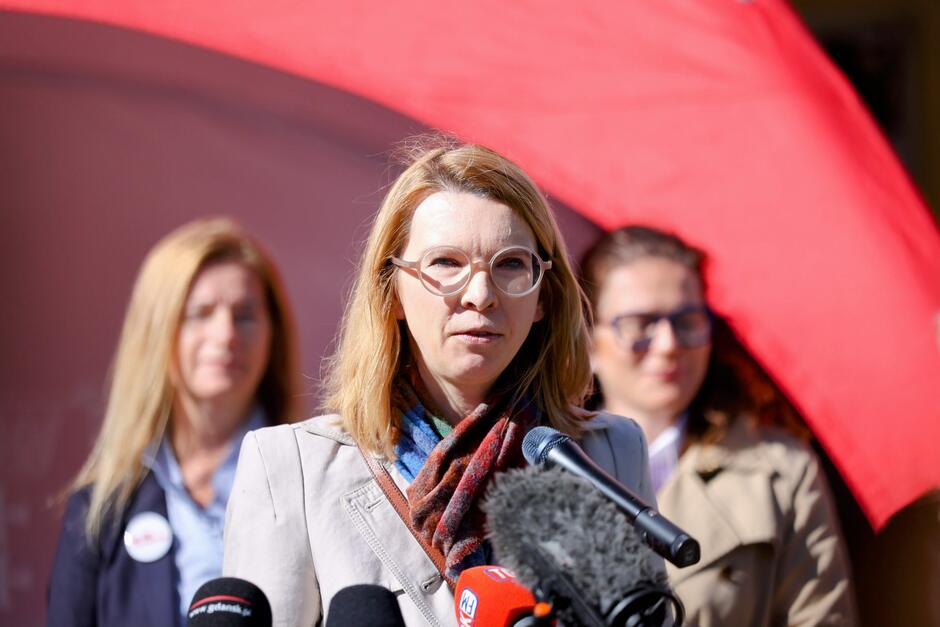 Na tym zdjęciu widzimy kolejną kobietę przemawiającą podczas tej samej konferencji prasowej, której tematem — jak wynika z wcześniejszych zdjęć — są nadchodzące wybory prezydenckie w Polsce 18 maja 2025 roku oraz zachęta do udziału w głosowaniu, szczególnie wśród studentów. Kobieta w jasnym płaszczu i kolorowym szalu stoi przed mikrofonami różnych mediów, m.in. RMF FM, Radio Gdańsk i TVN24. Za nią w tle stoją inne uczestniczki wydarzenia – kobiety widoczne wcześniej, w tym ta w beżowym płaszczu i niebieskich okularach.