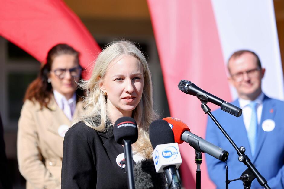 Na zdjęciu widzimy młodą kobietę z blond włosami przemawiającą do mediów podczas oficjalnego wydarzenia. Stoi przed kilkoma mikrofonami, m.in. stacji TVN24, Radio Gdańsk i innych. Ma na sobie czarną marynarkę z przypinką — podobną do tych, które nosili pozostali uczestnicy wydarzenia, co wskazuje na wspólną akcję informacyjną lub kampanijną. W tle widzimy osoby znane z wcześniejszych zdjęć — kobietę w beżowym płaszczu oraz mężczyznę w niebieskim garniturze, co potwierdza, że wszystkie fotografie pochodzą z tego samego wydarzenia. Tło stanowią biało-czerwone elementy (flagi, banery), które podkreślają patriotyczny i obywatelski charakter wydarzenia, najpewniej związanego z promocją udziału w wyborach prezydenckich 2025.