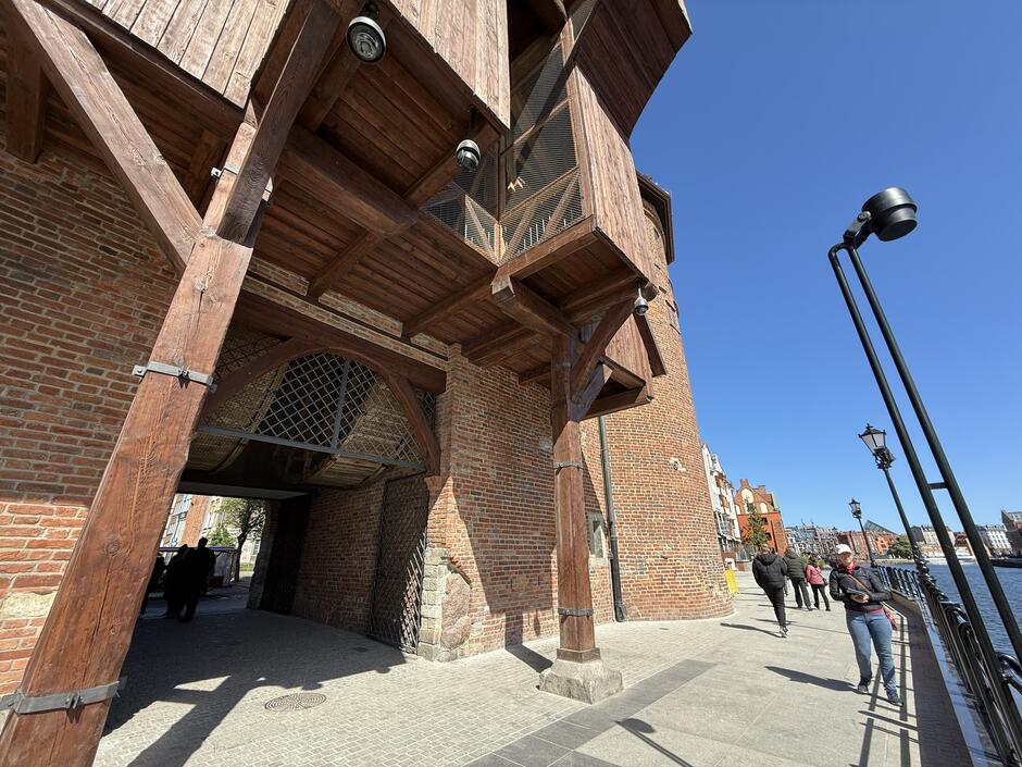 Fotografia ukazuje zewnętrzną stronę tej samej bramy, z drewnianą konstrukcją i detalami architektonicznymi nad wejściem. W tle widać deptak nad rzeką, ludzi spacerujących oraz charakterystyczne, kolorowe kamienice Gdańska.