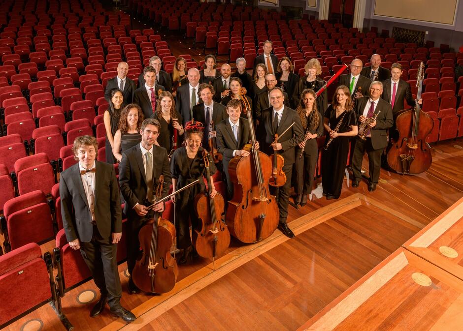 Na zdjęciu widzimy Scottish Chamber Orchestra – zespół muzyków pozujących wspólnie na tle pustej sali koncertowej, gotowych do występu