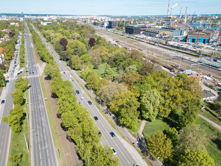 Zdjęcie lotnicze ukazuje szeroką aleję z torami tramwajowymi pośrodku, biegnącą równolegle do linii kolejowej oraz terenu przemysłowego z halami i kominami w tle. Po lewej stronie widoczny jest gęsto zalesiony park, a po prawej – infrastruktura kolejowa oraz zabudowania portowe, co sugeruje, że zdjęcie mogło zostać wykonane w Gdyni lub Gdańsku.