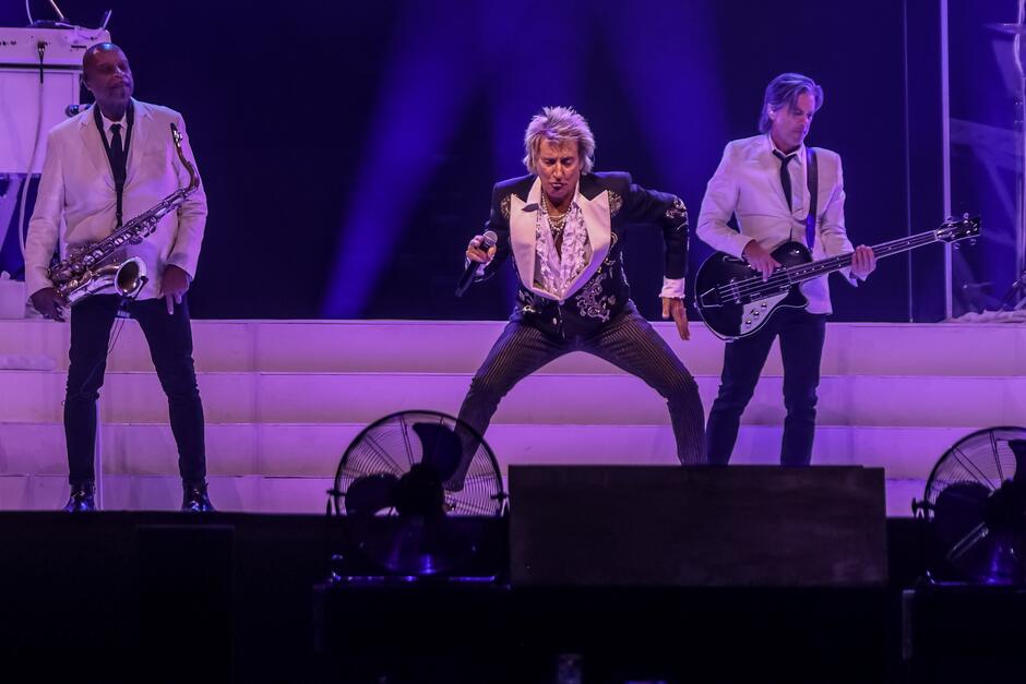rod stewart tańczący na scenie, za nim stoją dwaj gitarzyści