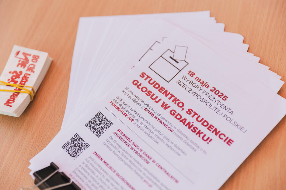 stos białych papierów z czerwonym tekstem i kodem QR na nich tekst na papierach wydaje się być zaproszeniem lub broszurą na ucznia ceremonii ukończenia studiów na górnym lewym rogu jest małe białe pudełko z czerwono-żółtym wzorem na nim obok pudełka, wydaje sie, że jest kilka innych przedmiotów na drewnianej powierzchni.