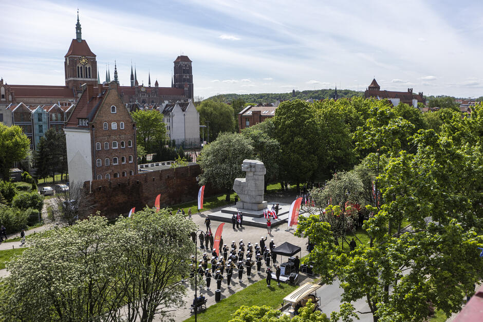 Widok z lotu ptaka na plac miejski z pomnikiem, pod którym odbywa się uroczystość patriotyczna. W tle widać zabytkowe, ceglane budynki. Wokoło rosną drzewa.