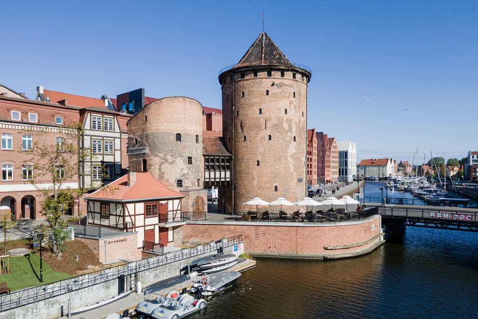  piękny widok na port z kilkoma łódkami zacumowanymi na wodzie. W środku znajduje się wysoka wieża ze spiczastym dachem i zegarkiem na górze. Wieża wykonana jest z kamienia i ma czerwoną ceglaną zewnętrzną. Znajduje się na moście nad rzeką, a po obu stronach znajdują się kilka mniejszych budynków. Po lewej i prawej stronie tej wieży widzimy rząd czerwonych domów z białymi dachami. Jest też kilka białych parasoli i stołów ustawionych wzdłuż nabrzeża. Niebo jest niebieskie, i pogoda wydaje się słoneczna i przejrzysta, co sugeruje, że zdjęcie zostało zrobione w ciągu dnia.