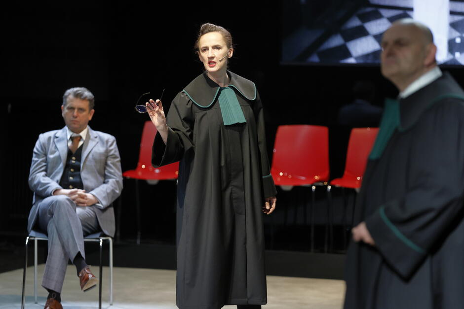 Kobieta w todze prawniczej z zielonymi akcentami gestykuluje podczas przemowy, trzymając okulary w jednej ręce — prawdopodobnie odgrywa rolę prokuratorki lub adwokatki. W tle widoczni są dwaj mężczyźni, również w togach, oraz czerwone krzesła