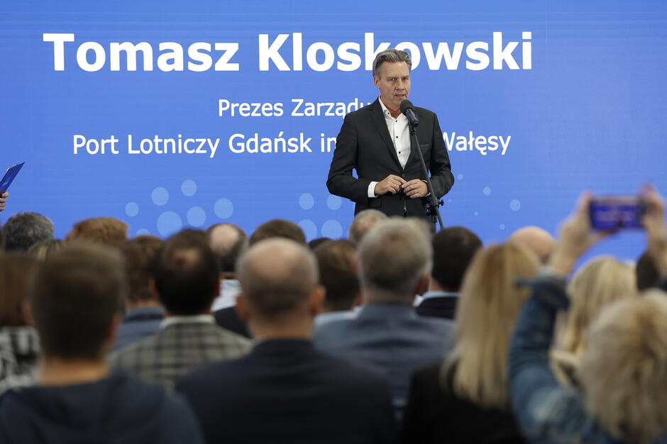Zdjęcie przedstawia Tomasza Kloskowskiego, prezesa zarządu Portu Lotniczego Gdańsk im. Lecha Wałęsy, przemawiającego do licznie zgromadzonej publiczności na tle niebieskiego ekranu z jego imieniem i funkcją. Atmosfera wydarzenia sugeruje oficjalną prezentację lub konferencję, dokumentowaną także przez uczestników.