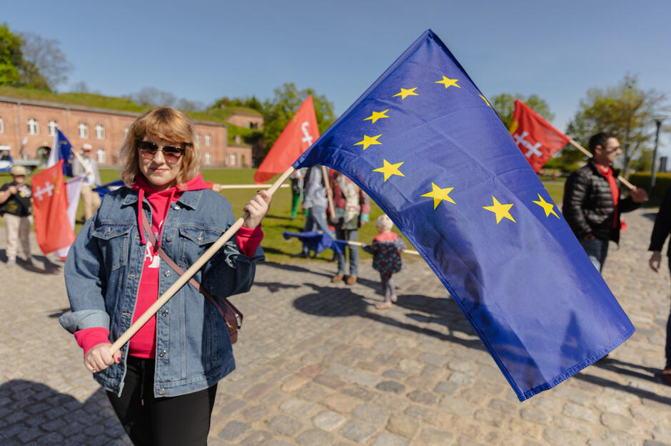  kobieta trzymająca dużą niebieską flagę z żółtymi gwiazdami, stoi na dziedzińcu z ceglanym budynkiem w tle, kobieta ma na szyi dżinsową kurtkę, okulary przeciwsłoneczne i czerwony szalik, za nią kilka innych osób trzymających czerwone flagi, z których niektóre wydają się być częścią protestu lub wiecu, na ziemi są drzewa i trawa, co sugeruje, że zdjęcie zostało zrobione w ciągu dnia.