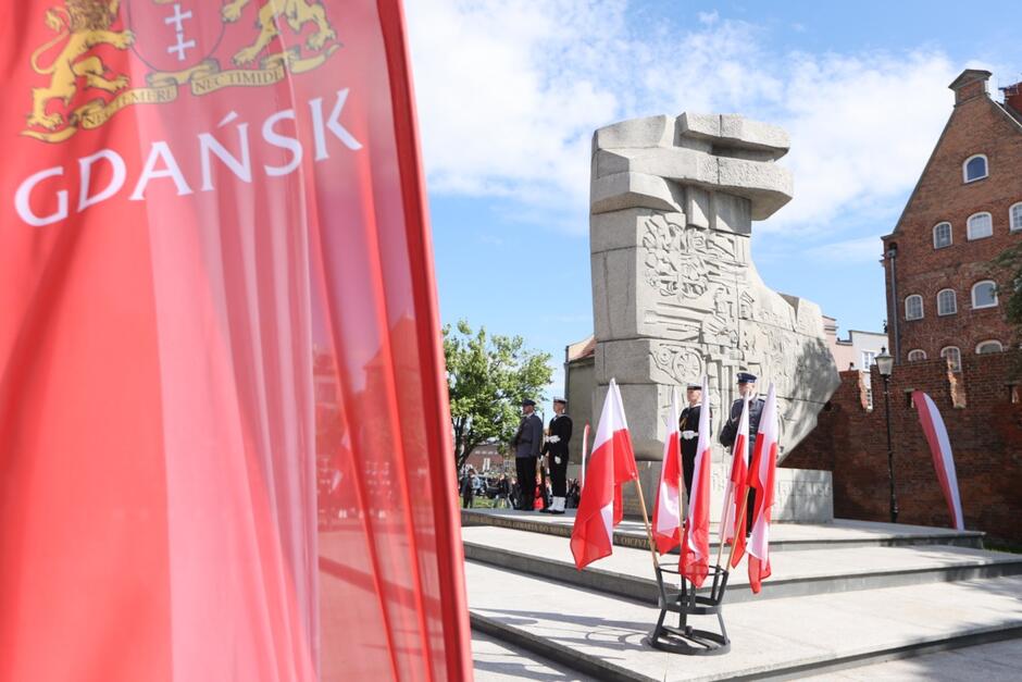 Na zdjęciu widzimy uroczystość patriotyczną odbywającą się w Gdańsku, przy Pomniku Obrońców Poczty Polskiej. Na pierwszym planie znajduje się czerwona flaga z herbem miasta Gdańska i jego nazwą. W tle widać pomnik wykonany z szarego kamienia, przy którym stoi warta honorowa – żołnierze lub funkcjonariusze w mundurach. Przed pomnikiem umieszczono kilka biało-czerwonych flag Polski, co podkreśla podniosły, narodowy charakter wydarzenia. Pogoda jest słoneczna, niebo prawie bezchmurne, co nadaje całości uroczysty nastrój. W tle widoczne są zabytkowe budynki i zebrani uczestnicy.
