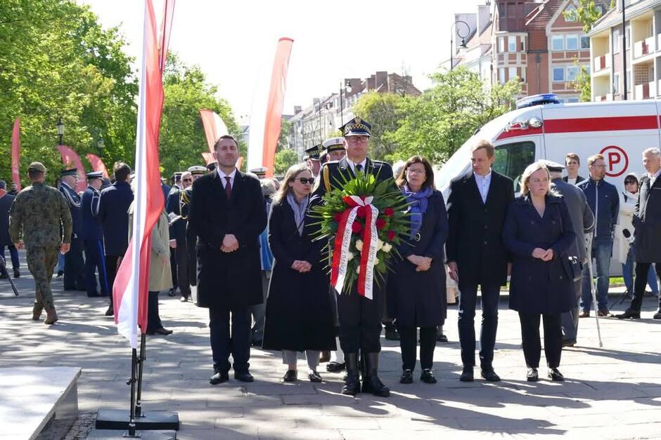 Na zdjęciu widzimy oficjalną delegację uczestniczącą w uroczystościach patriotycznych, prawdopodobnie związanych z rocznicą ważnego wydarzenia historycznego. W centrum znajduje się funkcjonariusz w mundurze galowym, trzymający wieniec z biało-czerwonymi wstążkami — symbol pamięci i hołdu. Po jego bokach stoją przedstawiciele władz samorządowych i instytucji, elegancko ubrani, w poważnym, skupionym nastroju. W tle widoczne są flagi Polski, uczestnicy uroczystości, żołnierze, funkcjonariusze oraz karetka pogotowia. Całość odbywa się na otwartym terenie miejskim w słoneczny dzień. Scena ta ukazuje powagę i szacunek wobec historii oraz bohaterów narodowych.