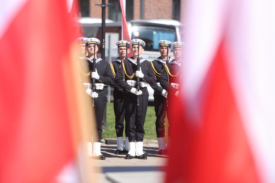 Na zdjęciu widzimy żołnierzy Marynarki Wojennej RP stojących w szyku honorowym podczas uroczystości państwowej lub wojskowej. Ubrani są w galowe mundury marynarskie z charakterystycznymi białymi czapkami oraz białymi rękawiczkami. Trzymają broń w postawie zasadniczej. Scena uchwycona została między biało-czerwonymi flagami Polski, co tworzy symboliczny i podniosły kadr, podkreślający patriotyczny charakter wydarzenia.
