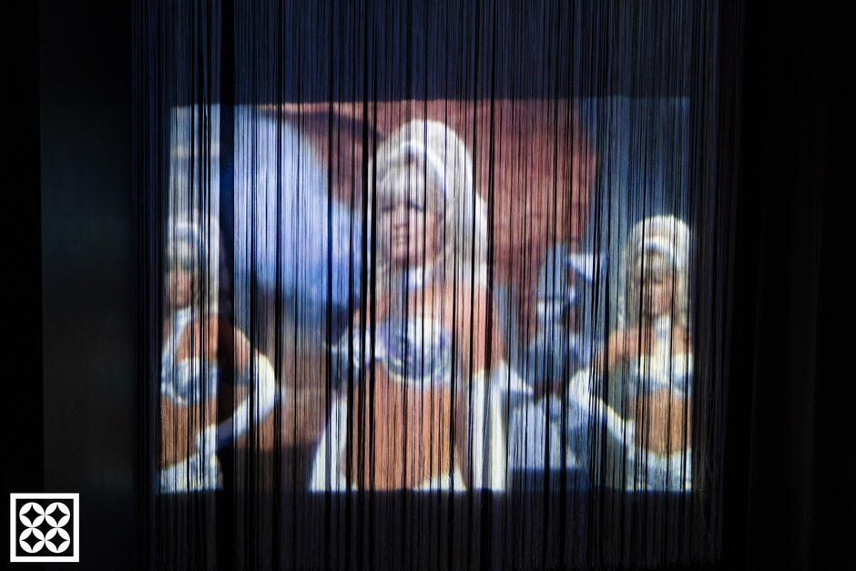 Kadr z filmu wyświetlany jest na cienkich, pionowo opadających niciach, które przypominają teatralną kurtynę, rozmywając i zniekształcając obraz. Trzy postacie w kostiumach retro tworzą surrealistyczną, niemal senno-futurystyczną scenę, która doskonale wpisuje się w eksperymentalny charakter wystawy