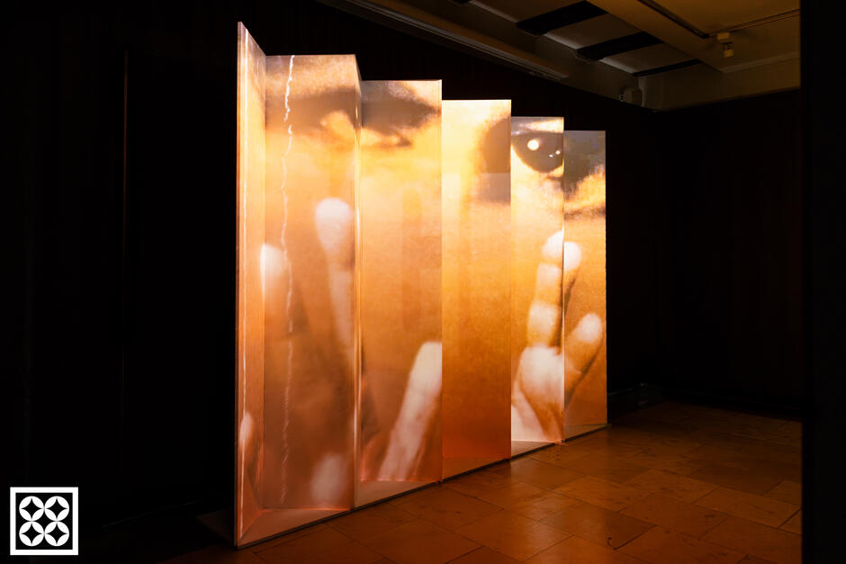 Zdjęcie przedstawia instalację artystyczną składającą się z kilku przezroczystych paneli ustawionych pod kątem, na których wyświetlony jest fragment powiększonego ludzkiego oblicza z widocznym okiem i dłońmi. Całość oświetlona jest ciepłym, pomarańczowym światłem, tworząc nastrojową i intymną atmosferę