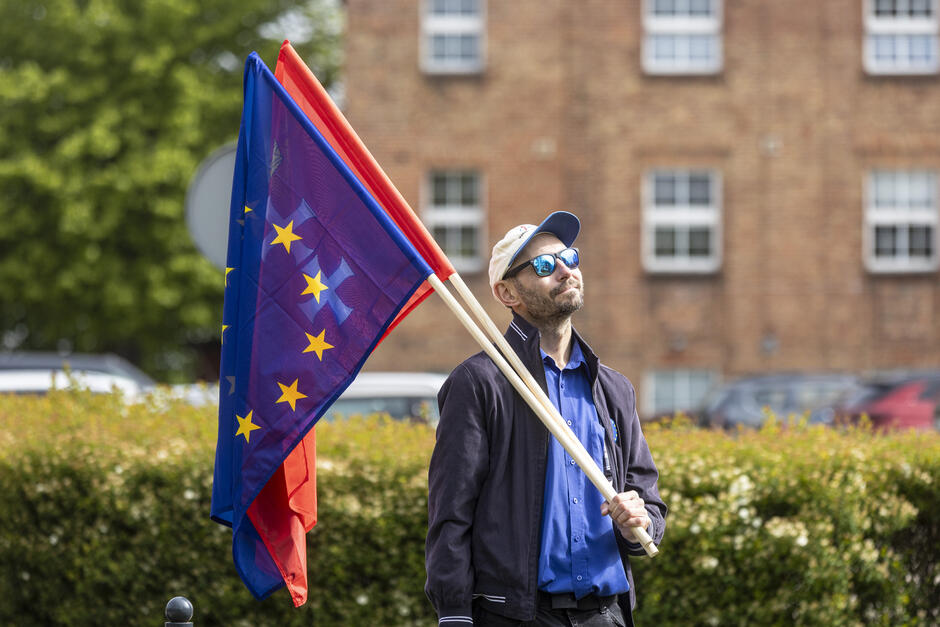 mężczyzna stojący przed ceglanym budynkiem, trzymający flagę z flagą Unii Europejskiej, posiadający niebieski płaszcz, białą czapkę i okulary przeciwsłoneczne, miał brodę i patrzy w niebo z poważnym wyrazem twarzy, za nim krzaki i drzewa, sugerujące, że znajduje się w dzielnicy mieszkalnej.