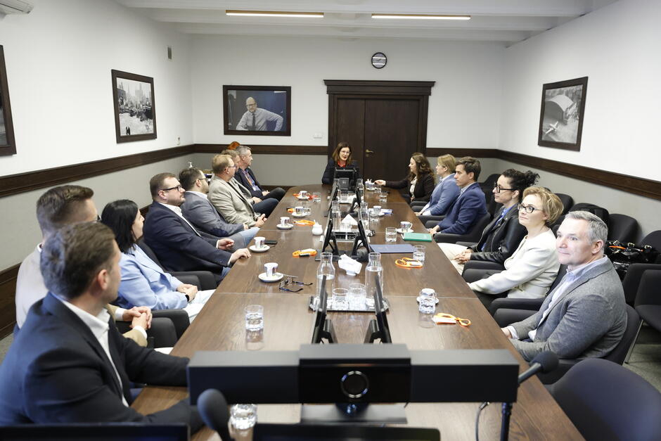 Kilkanaście osób siedzi wokół stołu w sali konferencyjnej.