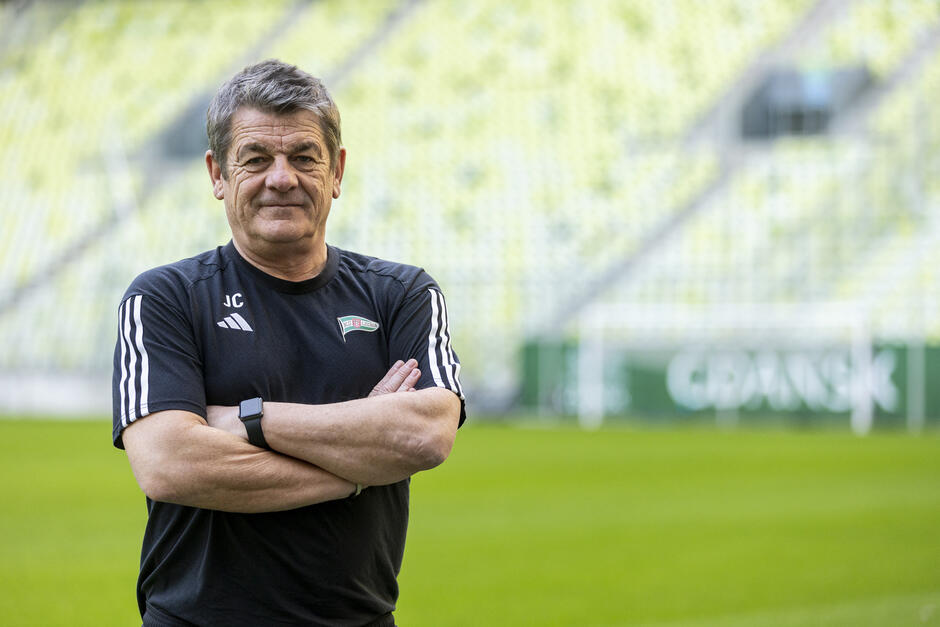 mężczyzna w średnim wieku stojący na boisku piłkarskim ze skrzyżowanymi rękami. Ma na sobie czarną koszulkę z logo drużyny sportowej i poważny wyraz twarzy. Tło jest zamazane, ale wydaje się, że stadion z pustymi miejscami i zielonym banerem. Mężczyzna jest prawdopodobnie trenerem lub menedżerem drużyny