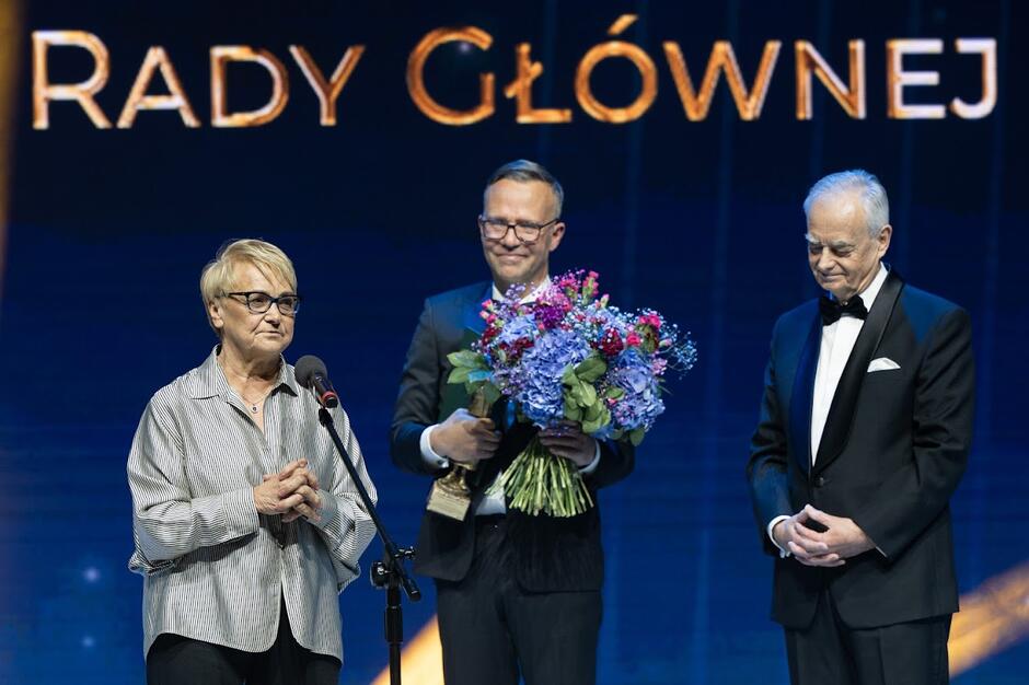 Trzy osoby na scenie podczas gali, kobieta po lewej, przemawia do mikrofonu