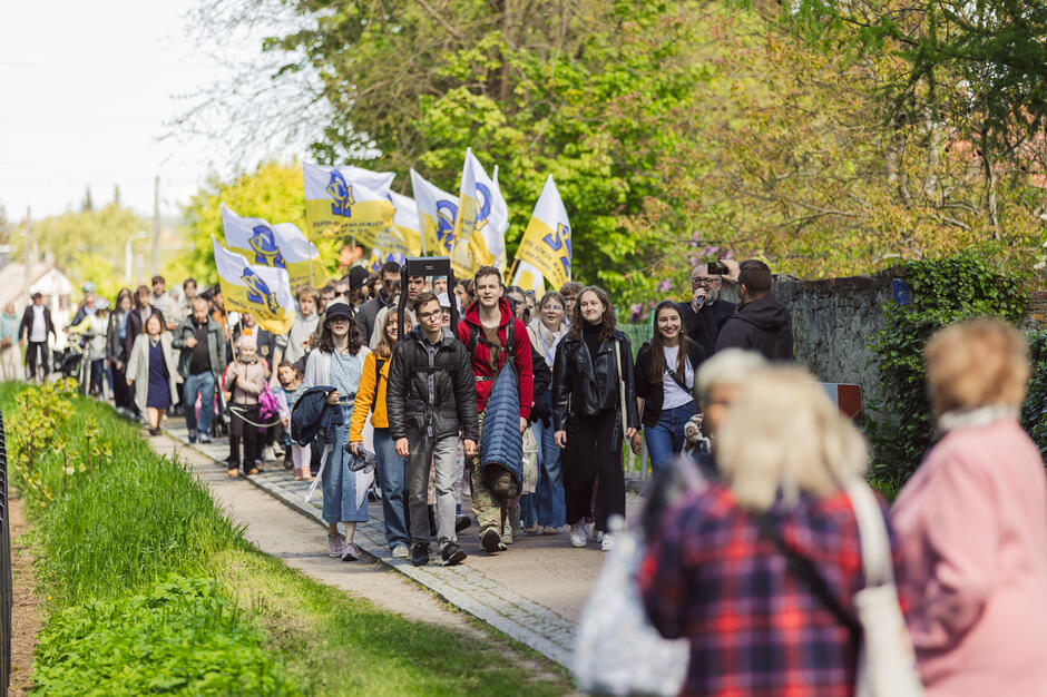 duża grupa ludzi spacerujących po ścieżce w parku. Ścieżka jest wyłożona drzewami i krzewami po obu stronach, a po prawej stronie kamienna ściana. W środku ścieżki jest kilka osób trzymających żółte i niebieskie flagi. Niektórzy ludzie chodzą plecami do kamery, podczas gdy inni noszą torby i inne przedmioty.