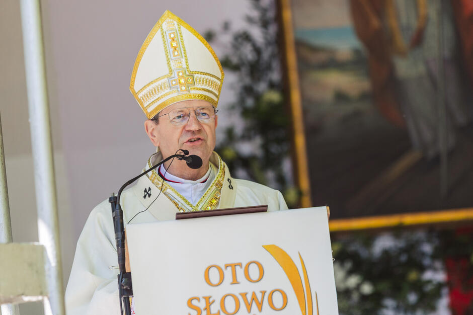 mężczyzna stojący za podium z mikrofonem przed sobą. Ma na sobie białą szatę kapłańską i żółto-złotą czapkę z napisanymi na niej słowami OTO SLOWO . Mężczyzna wydaje się wygłaszać przemówienie lub prezentację, ponieważ za nim stoi transparent z napisem Oto Slowo . Drzewa i kwiaty w tle sugerują, że obraz został zrobiony na wydarzeniu religijnym.
