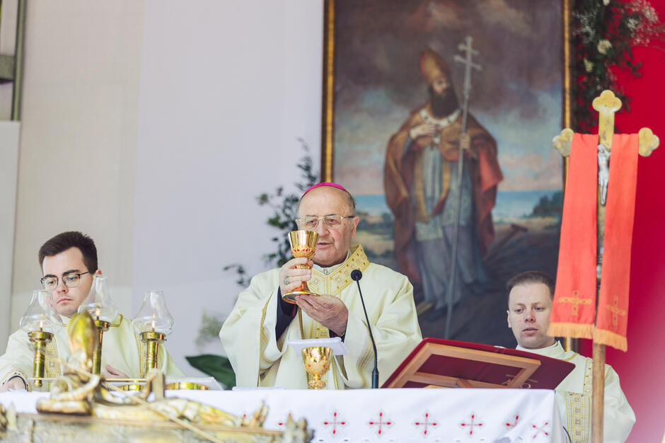 Kapłan w białej szacie i czerwonej czapce, trzymający złoty kielich i mówiący do mikrofonu; stoi za stołem z krzyżem i innymi przedmiotami religijnymi; po lewej stronie młody człowiek siedzi przy ołtarzu, a po prawej stronie klęka przed nim; w tle wisi na ścianie obraz mężczyzny o długich włosach i brodzie; kapłan wydaje się głosić kazanie lub błogosławieństwo ołu.