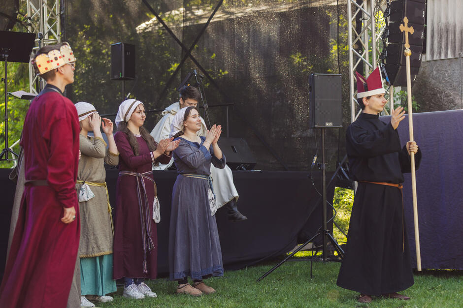 grupa ludzi stojących na scenie w parku lub ogrodzie, na zdjęciu sześć osób, trzech mężczyzn i trzy kobiety, wszystkie ubrane w stroje średniowieczne, mężczyzna po lewej stronie nosi czerwoną szatę i koronę, podczas gdy kobieta pośrodku klepie ręce, po prawej stronie mężczyźni ubrani w kapłana, trzymając sztabek z krzyżem, stoi przed mikrofonem i wydaje się wygłaszać przemówienie lub prezentację, w tle widzimy dużą metalową konstrukcję i drzewa.