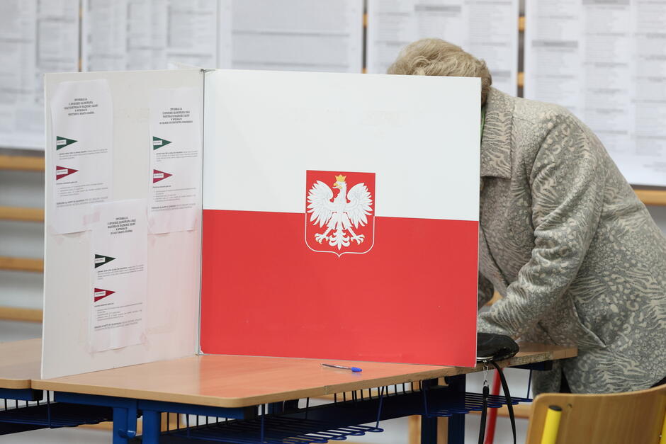 Zdjęcie przedstawia osobę głosującą w lokalu wyborczym w Polsce, zasłoniętą parawanem z godłem narodowym na tle biało-czerwonej flagi. Wnętrze lokalu wskazuje na oficjalną procedurę wyborczą, podkreślając wagę udziału obywateli w demokratycznym procesie.