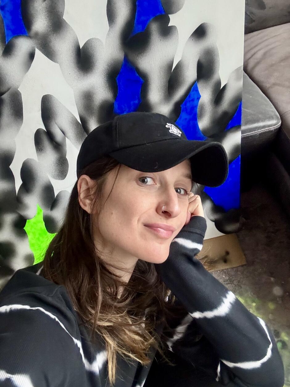 na zdjęciu znajduje się kobieta w czarnej czapce z daszkiem, na tle obrazu