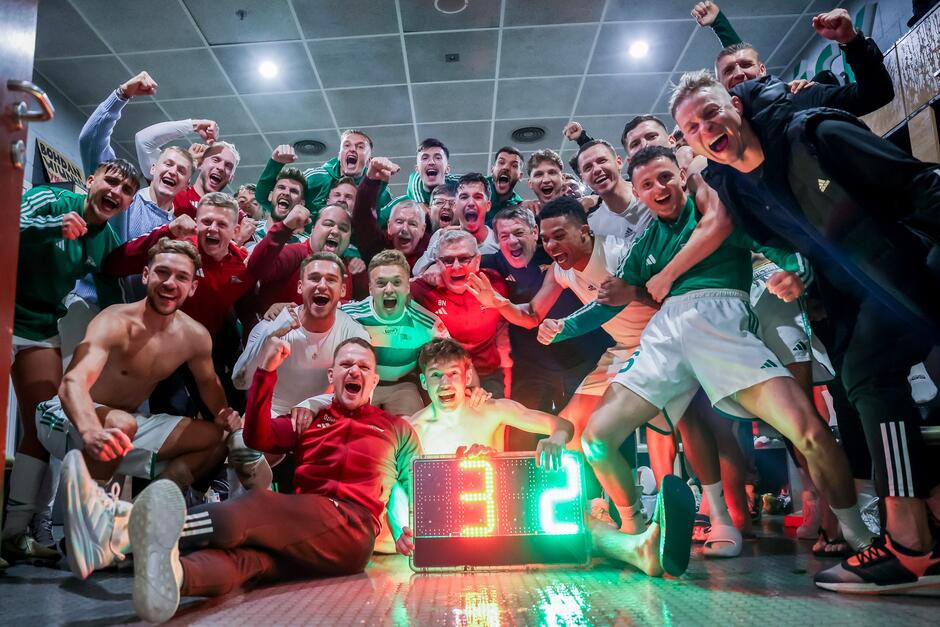 Grupa zawodników i sztabowców w szatni - pozują do wspólnego zdjęcia, entuzjastyczne uśmiechy i gesty