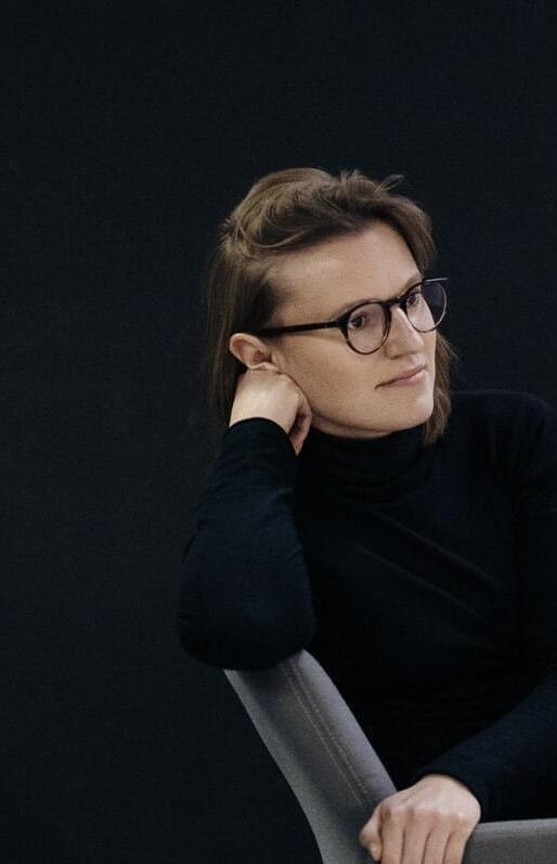 Na zdjęciu młoda kobieta w czarnej koszulce, w okularach, siedząca bokiem na krześle.