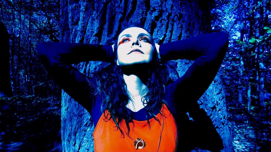 Na zdjęciu kobieta w pomarańczowej koszulce, z czarnymi rękawami, z założonymi rękoma na uszy. W tle drzewo.